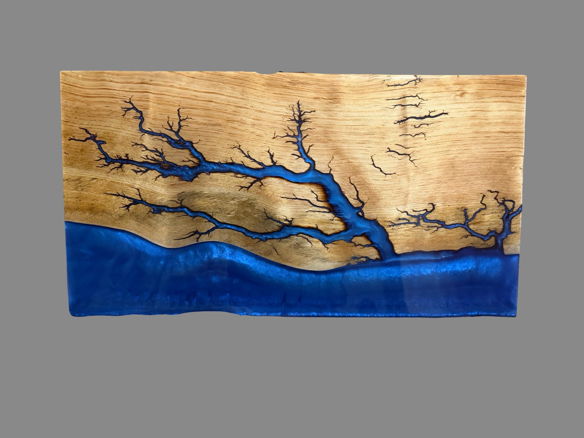 A rectangular wood plaque featuring a natural live-edge wood grain background with a bright blue resin river running through it, resembling a tree branch or canyon landscape, shown against a solid gray background.